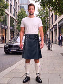 A man stands on a city sidewalk wearing a white crew-neck t-shirt and a dark gray pleated kilt with silver metal accents. He is also wearing off-white knee-high socks and black leather shoes.