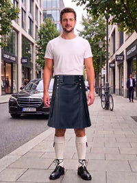 A man stands on a city sidewalk wearing a white crew-neck t-shirt and a dark gray pleated kilt with silver metal accents. He is also wearing off-white knee-high socks and black leather shoes.