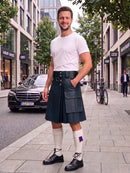 A man standing on a city sidewalk wearing a dark green pleated kilt, white t-shirt, white knee-high socks, and black leather shoes. A black Mercedes-Benz car is parked on the street behind him.