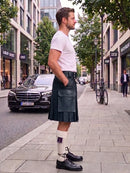 A man standing on a city sidewalk wearing a dark green leather kilt, white t-shirt, white knee-high socks with a purple ribbon, and black leather shoes. A black Mercedes-Benz car and a bicycle are parked on the street behind him.