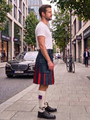 A man standing on a city sidewalk wearing a traditional Scottish kilt with red and dark blue stripes, a white t-shirt, white socks, and black leather shoes. A black Mercedes-Benz car is parked on the street behind him.