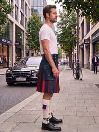 A man standing on a city sidewalk wearing a traditional Scottish kilt with red and dark blue stripes, a white t-shirt, white socks, and black leather shoes. A black Mercedes-Benz car is parked on the street behind him.