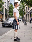 A man stands on a city sidewalk wearing a white short-sleeve T-shirt, a navy kilt with white vertical stripes and white pocket detailing, cream knee-high socks, and black leather oxford shoes. Buildings, trees, a parked black car, and a bicycle are visible in the urban street setting.
