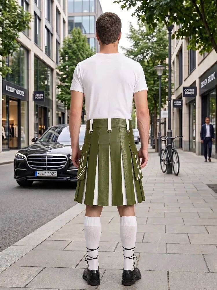 A man stands on a city sidewalk facing away from the camera, wearing an olive green pleated kilt with white vertical stripe panels, a white t-shirt, white knee-high socks, and black shoes. Storefronts, trees, and parked cars are visible in the background.