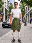 A man stands on a city sidewalk wearing a white crew-neck t-shirt and an olive green kilt with white geometric stripe patterns. He is also wearing white knee-high socks and black leather shoes.