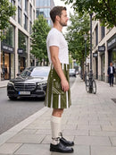 A man stands on a city sidewalk wearing a green and white striped kilt with a white t-shirt, cream knee-high socks, and black leather shoes. A black Mercedes-Benz car and a bicycle are parked on the street behind him.