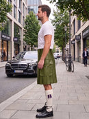 A man standing on a city sidewalk wearing a brown leather kilt, white t-shirt, white socks, and black shoes, with a street scene in the background.