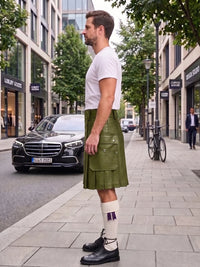 A man standing on a city sidewalk wearing a brown leather kilt, white t-shirt, white socks, and black shoes, with a street scene in the background.