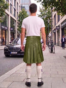 A man standing on a city sidewalk, viewed from behind, wearing an olive green leather pleated kilt, a white t-shirt, white knee-high socks, and black shoes. Buildings, trees, a black Mercedes, and a bicycle are visible in the background.