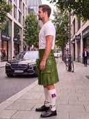 A man standing on a city sidewalk wearing a white t-shirt, an olive green kilt with multiple utility pockets, white knee-high socks with a purple accent, and black leather shoes. A black Mercedes-Benz car is parked on the street behind him.