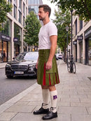 A man standing on a city sidewalk wearing a green pleated leather kilt with red and purple striped accents, a white t-shirt, white knee-high socks, and black leather shoes. A black Mercedes-Benz is parked on the street behind him.