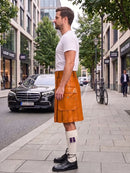 A man standing on a city sidewalk wearing a bright orange-brown leather kilt, a white t-shirt, white socks, and black leather shoes. A black Mercedes-Benz car is parked on the street behind him, with trees and storefronts in the background.