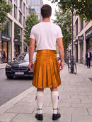 A man standing on a city sidewalk, viewed from behind, wearing a white t-shirt and a mustard-yellow leather kilt with side buckles, white knee-high socks, and black shoes. Buildings, trees, a black Mercedes, and a bicycle are visible in the background.