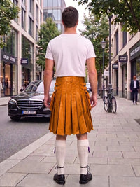 A man standing on a city sidewalk, viewed from behind, wearing a white t-shirt and a mustard-yellow leather kilt with side buckles, white knee-high socks, and black shoes. Buildings, trees, a black Mercedes, and a bicycle are visible in the background.