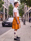 A man standing on a city sidewalk wearing a bright orange leather kilt, a white t-shirt, white socks, and black leather shoes. A black Mercedes-Benz car is parked on the street behind him, with trees and buildings in the background.