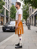 A man standing on a city sidewalk wearing a bright orange and white pleated kilt, a white t-shirt, white knee-high socks, and black leather shoes. A black Mercedes-Benz car is parked on the street behind him, with trees and storefronts in the background.