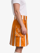 A side view of a person wearing a bright orange pleated kilt with white vertical stripes and a large front flap pocket, paired with a white t-shirt against a light gray background.