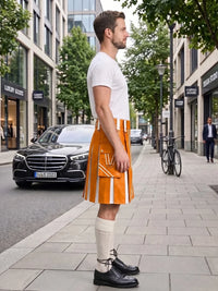 A man stands on a city sidewalk wearing a bright orange kilt with white vertical stripe details, a white t-shirt, cream knee-high socks, and black leather dress shoes. Modern buildings, trees, a parked black Mercedes, and a bicycle are visible in the background.