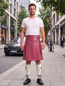 A man stands on a city sidewalk wearing a pink faux-leather kilt, a white t-shirt, white knee-high socks, and black leather shoes. The background shows a street with buildings, a black Mercedes-Benz car, and a bicycle.