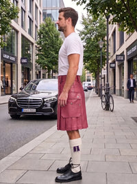 A man standing on a city sidewalk wearing a white t-shirt, a dusty rose pink faux-leather kilt with silver zippers, white socks with purple accents, and black leather shoes. A black Mercedes-Benz is parked in the street behind him.