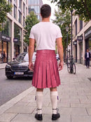 A man viewed from behind wearing a white t-shirt and a pink pleated faux-leather kilt, standing on a city sidewalk next to a street with a Mercedes-Benz car and trees.