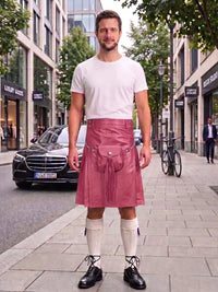 A man standing on a city sidewalk wearing a pink faux-leather kilt with a front pouch and drawstring, paired with a white t-shirt, white knee-high socks, and black leather shoes. A black Mercedes-Benz is parked on the street behind him.