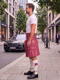 A man standing on a city sidewalk wearing a pink faux-leather kilt, white t-shirt, white knee-high socks, and black shoes, with a street scene and parked cars in the background.