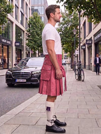 A man standing on a city sidewalk wearing a pink leather kilt, white t-shirt, white socks, and black leather shoes, with a black Mercedes and urban buildings in the background.