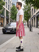 A man standing on a city sidewalk wearing a pink and white striped kilt, a white t-shirt, white knee-high socks, and black leather shoes, with a street and storefronts in the background.