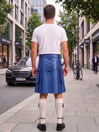 A man standing on a city sidewalk, viewed from behind, wearing a blue kilt with lace-up back detailing, a white t-shirt, white knee-high socks, and black shoes. Storefronts, trees, and parked cars are visible in the background.