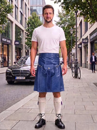 A man stands on a city sidewalk wearing a white crew-neck t-shirt and a blue leather kilt with a waist strap, paired with cream knee-high socks and black leather shoes. A black Mercedes-Benz car and a bicycle are visible in the background of the street scene.