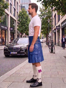 A man standing on a city sidewalk wearing a blue leather kilt, white t-shirt, white socks, and black shoes, with a black car and urban buildings in the background.