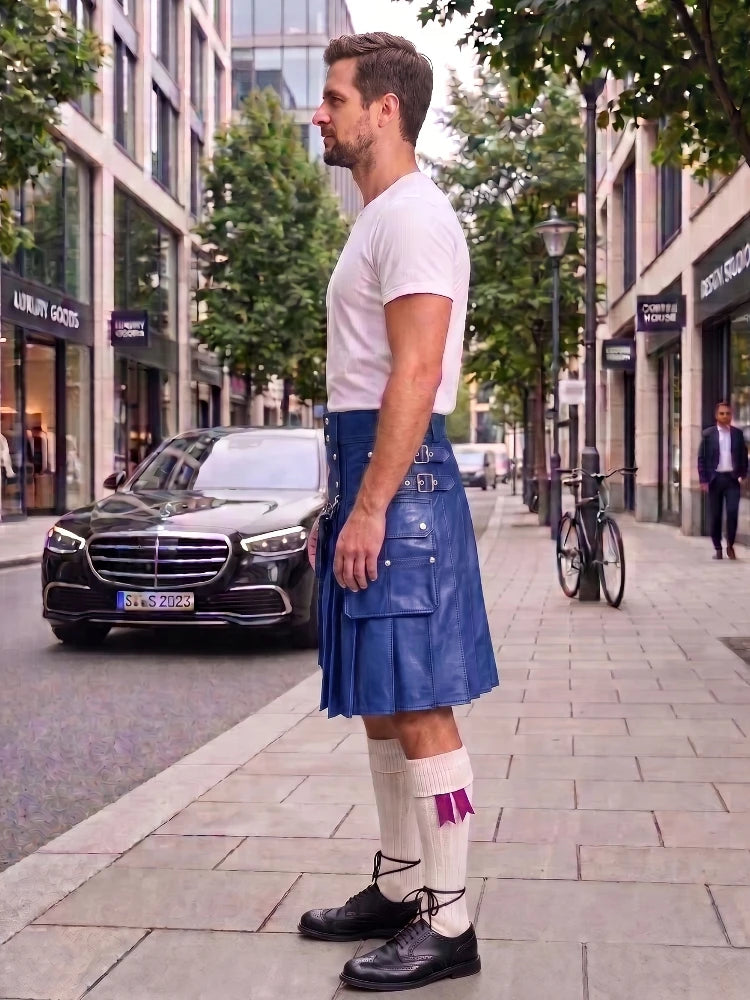 A man standing on a city sidewalk wearing a blue leather kilt, white t-shirt, white socks, and black shoes, with a black car and urban buildings in the background.