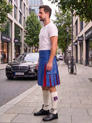 A man stands on a city sidewalk wearing a blue leather kilt with red and purple pleated accents, a white t-shirt, white knee-high socks, and black shoes. Buildings, trees, a parked black car, and a bicycle are visible in the background.