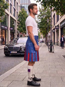 A man standing on a city sidewalk wearing a blue and red pleated kilt, a white t-shirt, white socks, and black leather shoes, with a street scene in the background.
