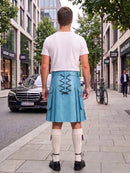 A man standing on a city sidewalk, photographed from behind, wearing a light blue pleated kilt with black lace-up detailing at the back, a white t-shirt, white knee-high socks, and black shoes. Buildings, trees, a black Mercedes, and a bicycle are visible in the background.