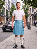 A man stands on a city sidewalk wearing a light blue pleated kilt with silver buttons, a white t-shirt, white knee-high socks, and black shoes. Buildings, trees, and parked cars are visible in the background.