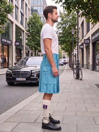 A man standing on a city sidewalk wearing a light blue pleated kilt, white t-shirt, white socks with purple stripes, and black leather shoes. A black Mercedes-Benz car is parked on the street behind him.