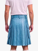 A person shown from the back wearing a light blue pleated leather kilt with side cargo pockets, paired with a pale pink t-shirt and white socks, against a light gray studio background.