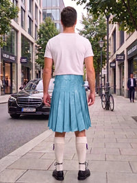 A man standing on a city sidewalk, viewed from behind, wearing a white t-shirt, a bright turquoise pleated leather kilt, white knee-high socks, and black leather shoes. Storefronts, trees, and a parked black Mercedes are visible in the background.