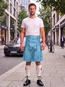 A man stands on a city sidewalk wearing a light blue denim kilt over a white t-shirt, cream knee-high socks, and black leather shoes. A black Mercedes-Benz is parked behind him on the street, with storefronts and trees in the background.