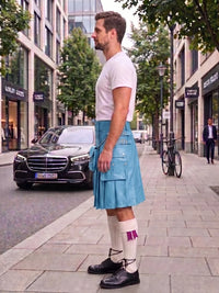 A man standing on a city sidewalk wearing a light blue kilt, white t-shirt, white knee-high socks, and black shoes, with buildings, trees, and a parked black car in the background.