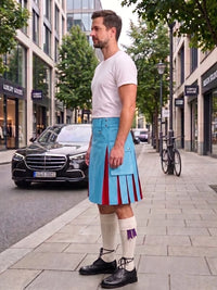 A man stands on a city sidewalk wearing a bright blue pleated kilt with red and purple stripe details, a white t-shirt, white knee-high socks, and black shoes. Cars and storefronts are visible in the background.