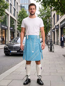 A man stands on a city sidewalk wearing a light blue kilt with white geometric line patterns, a white crew-neck t-shirt, white knee-high socks, and black leather shoes. The background shows a blurred street scene with buildings, a parked black car, and a bicycle.