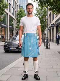 A man stands on a city sidewalk wearing a light blue kilt with white geometric line patterns, a white crew-neck t-shirt, white knee-high socks, and black leather shoes. The background shows a blurred street scene with buildings, a parked black car, and a bicycle.