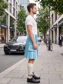 A man standing on a city sidewalk wearing a white t-shirt, a light blue pleated kilt with white vertical stripes and flap pockets, cream knee-high socks, and black leather shoes. A black Mercedes-Benz car and a bicycle are parked on the street behind him.