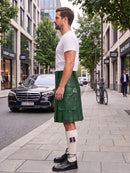 A man standing on a city sidewalk wearing a white crew-neck t-shirt, a dark gray pleated kilt with multiple front flap pockets, white knee-high socks with a purple accent, and black leather lace-up shoes. A black Mercedes-Benz car and a bicycle are parked on the street behind him.