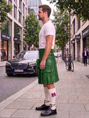 A man standing on a city sidewalk wearing a bright green leather kilt, a white t-shirt, white knee-high socks, and black leather shoes. A black Mercedes-Benz car is parked on the street behind him, with trees and storefronts in the background.