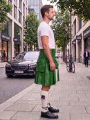 A man standing on a city sidewalk wearing a green leather kilt, white t-shirt, white socks, and black leather shoes, with a Mercedes-Benz and urban buildings in the background.