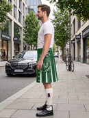 A man standing on a city sidewalk wearing a green and white kilt, white t-shirt, white socks, and black shoes, with a street scene and parked cars in the background.
